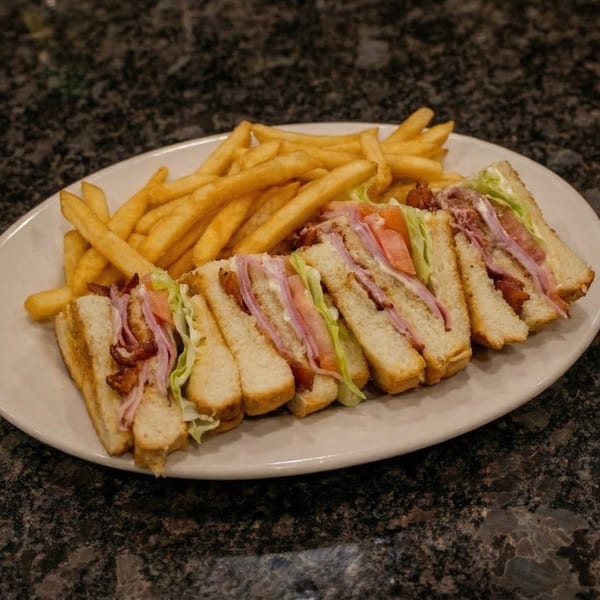 A club sandwich cut into four triangles with bacon, ham, lettuce, and tomato, served with fries.